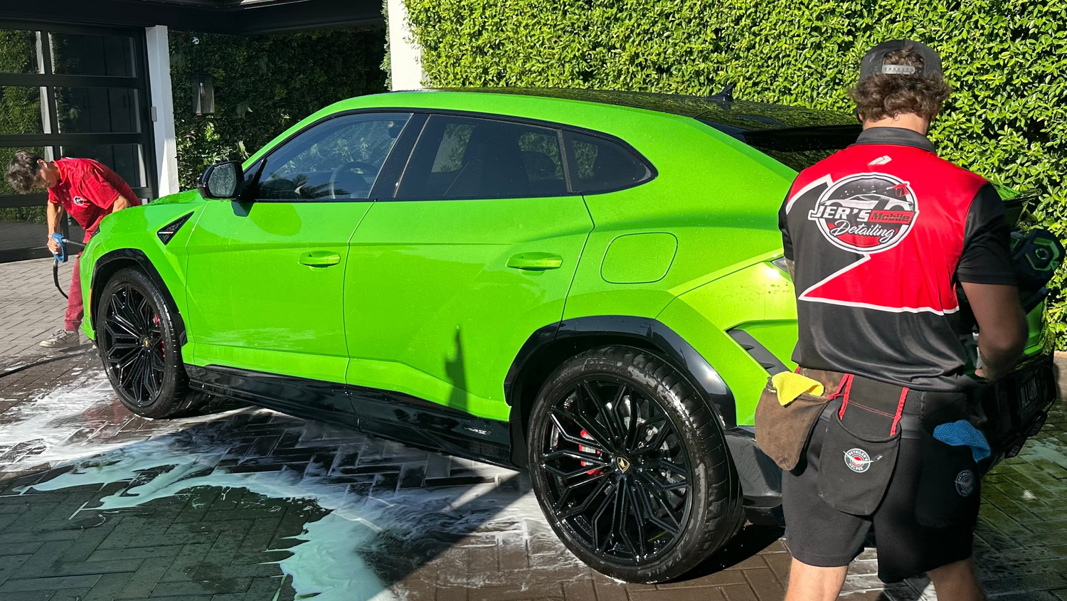 A bright lime green Lamborghini Urus SUV is being washed by hand in a driveway, with a worker in Jer's Mobile Detailing  uniform standing next to it against a green hedge background.