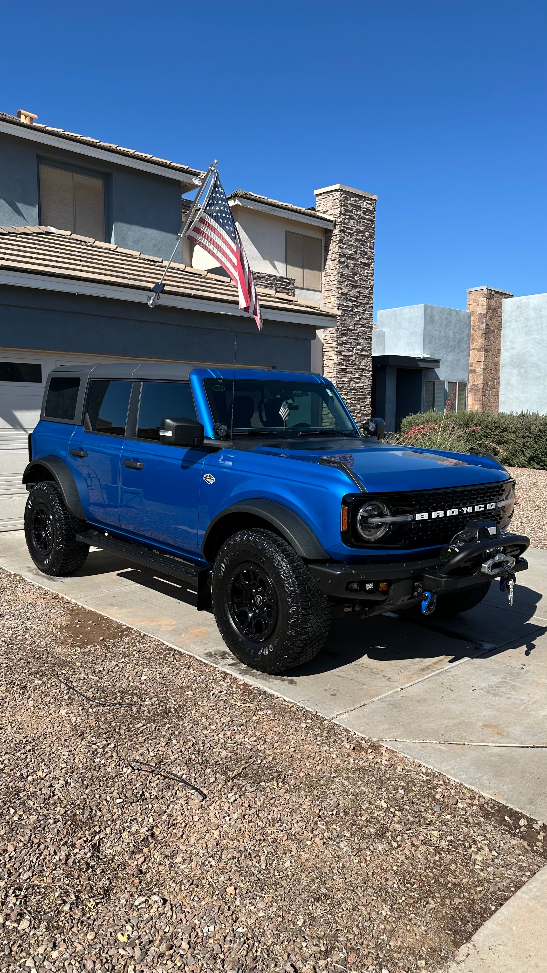 A bright blue Ford Bronco parked on a concrete driveway in front of a suburban house, with a flag flying from the garage and a clear blue sky above.
